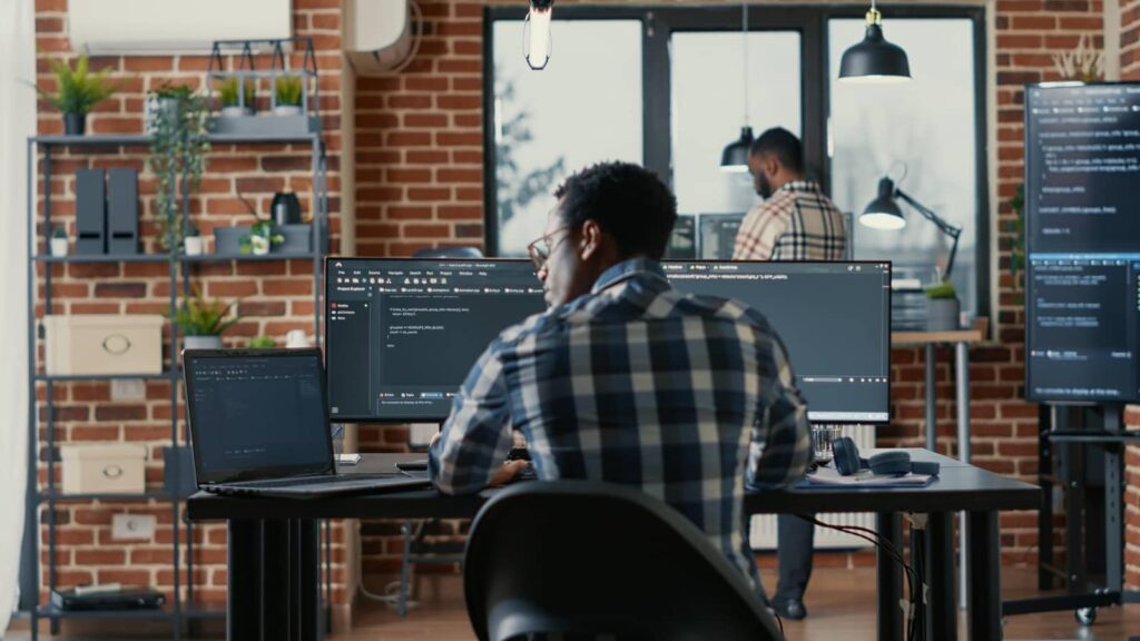 Programmer coding on laptop sitting down at desk with computer screens
