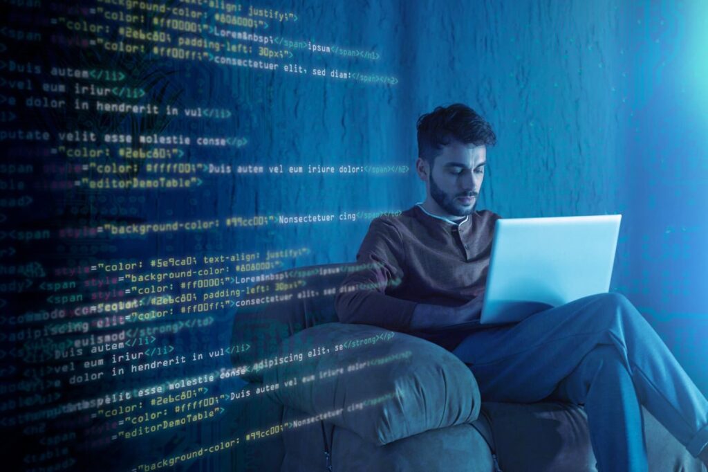 A man sits in a chair with a laptop, program code is on the wall next to him